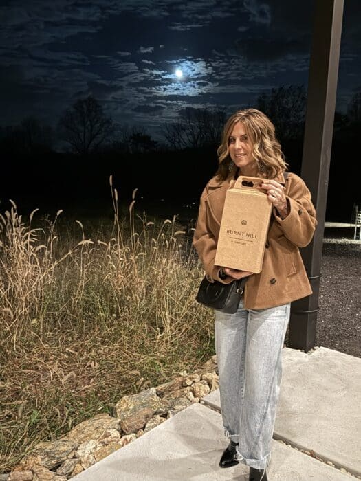 a Woman with Wavy Hair Stands Outdoors at Night Holding a Burnt Hill Wine Box from Burnt Hill Farm She Wears a Brown Jacket Light Jeans and Black Boots Behind Her Are Tall Grasses Rocks and a Dark Sky with a Full Moon Partially Veiled by Clouds