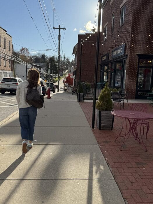 a Person with Shoulder Length Hair Wearing a Gray Top Blue Jeans and White Shoes Walks on a Sunny Sidewalk in Mt Airy Md Next to a Brick Building Red Metal Table and Chairs String Lights and Parked Cars Complete the Lively Streetscape