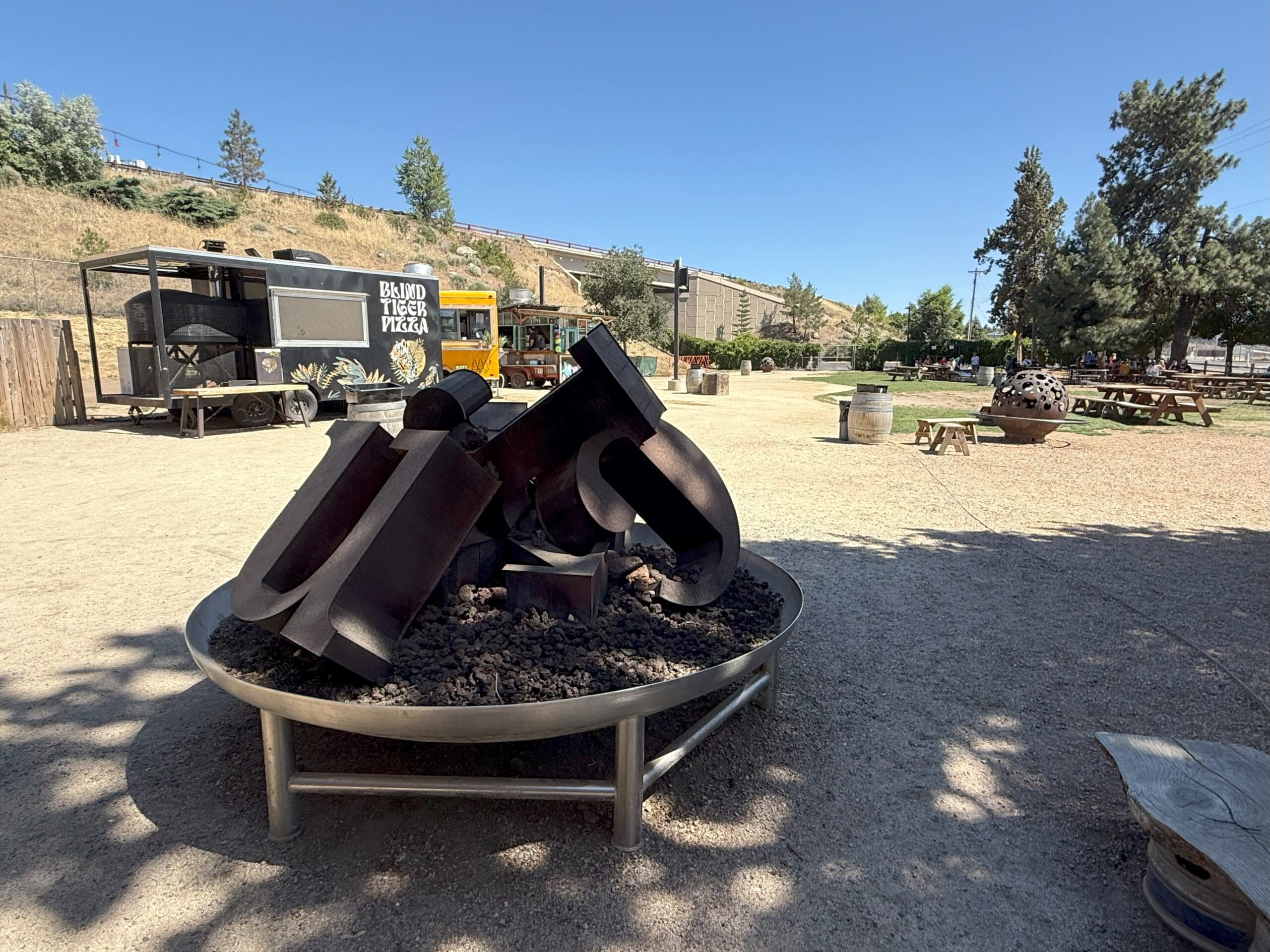 a Metal Sculpture of Large Abstract Letters Sits in a Circular Planter Filled with Dark Rocks on a Gravel Lot Near Picnic Tables at Crux Fermentation Project in the Background Theres a Food Truck Labeled bavid Beer Yoda Trees and a Clear Blue Sky