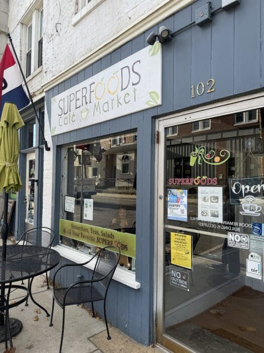 a Street View of Superfoods Café Market in Mt Airy Md Features a Blue Gray Exterior the Sign Advertises Smoothies Teas Salads and Superfoods an Outdoor Table Sits on the Sidewalk with Business Hours and a Wi Fi Sign in the Window