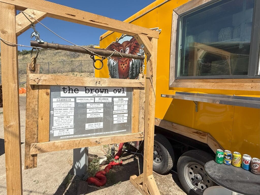 a Wooden Sign Reading the Brown Owl Displays a Menu in Front of a Yellow Food Truck a Painted Owl Decorates the Truckone of Many Unique Bend Oregon Restaurants Cans of Sodas Sit on a Round Gray Table Nearby with Sunlight and Dry Grass in the Background