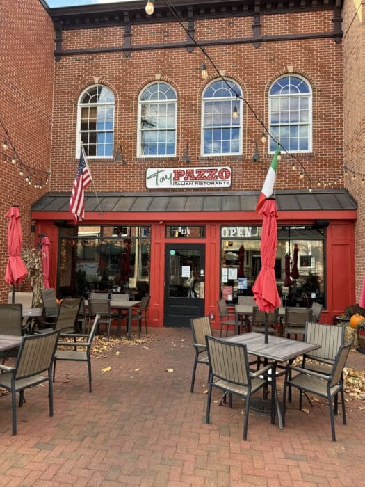 a Brick Italian Restaurant in Mt Airy Md with Large Arched Windows String Lights American and Italian Flags Red Umbrellas over Outdoor Tables and Chairs and a Sign Reading Pazzo Italian Ristorante Fallen Leaves Are Scattered on the Ground