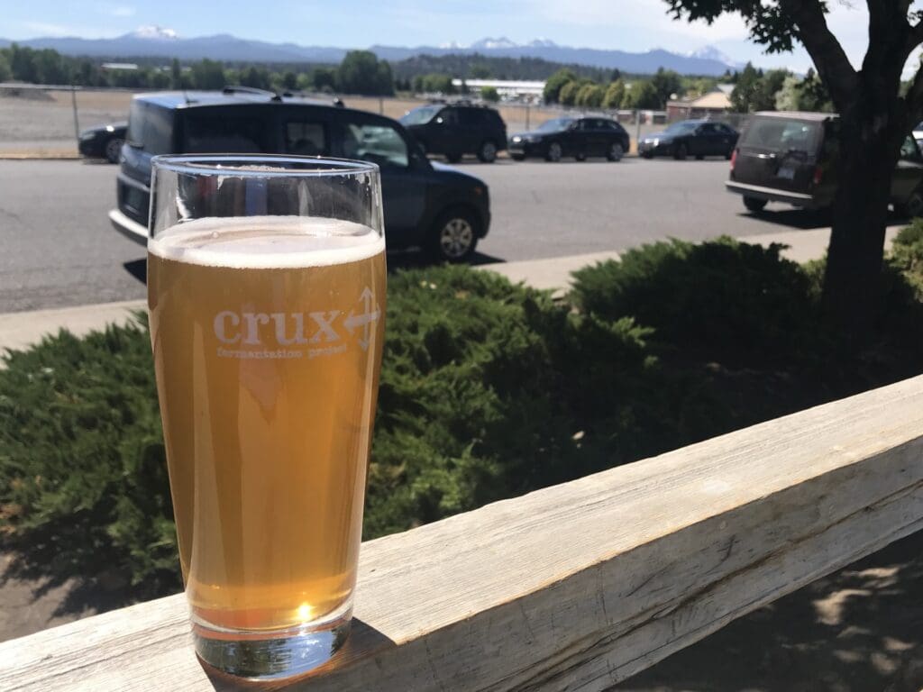 a Pint Glass of Hazy Beer Branded with Crux Fermentation Project Rests on a Wooden Railing with a Road Parked Cars Lush Green Bushes and Distant Mountains Framed by a Clear Blue Sky in the Background