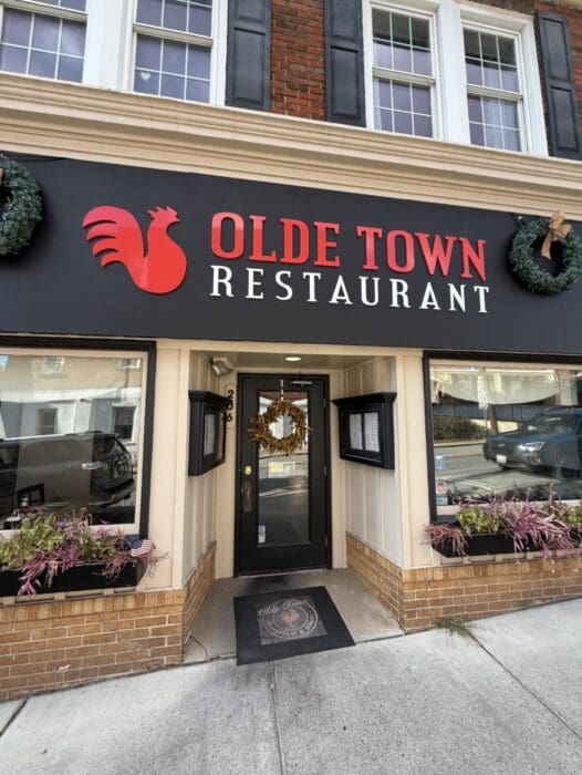 Storefront of Olde Town Restaurant in Mt Airy Md with a Black and Red Sign Featuring a Rooster Logo Above the Entrance the Door Has a Holiday Wreath and Window Boxes with Plants Sit Beneath Large Windows Framed by Brick and Beige Siding