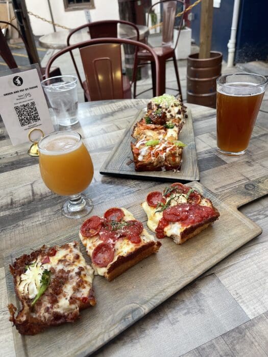 Two Wooden Boards Hold Detroit style Pizza Slices with Toppings Like Pepperoni Sausage Jalape&ntilde;o and Vegetables Nearby Two Beers and a Qr Code Menu Add to the Vibe of Enjoying Pizza in Fredrick Md at This Inviting Outdoor Dining Spot