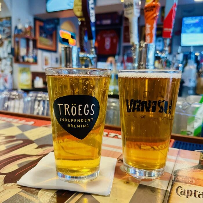 Two Pint Glasses of Golden Beer Sit on a Bar Counterone with a Tr&ouml;egs Independent Brewing Logo the Other Vanish Both Rest on White Napkins Behind Them Are Beer Taps and Blurred Lights Perfect for Enjoying After Pizza in Frederick Md