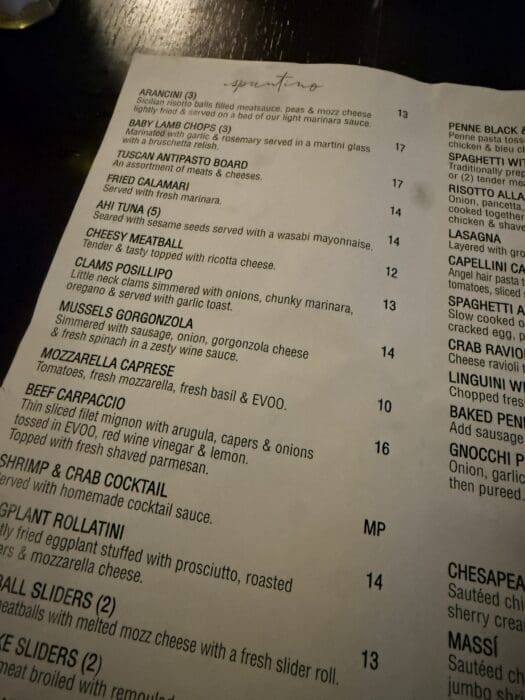 A close-up photo of a restaurant menu at Cucina Massi Frederick MD on a dark table, showing items like arancini, lamb chops, antipasto board, spicy calamari, meatballs, fried mozzarella, caprese, shrimp cocktail, rollatini, and sliders with prices listed.