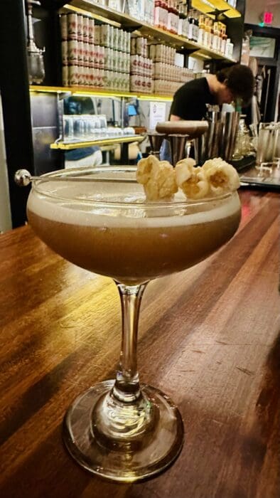 a Creamy Cocktail Garnished with Popped Popcorn Sits in a Coupe Glass on a Wooden Bar Counter at Cucina Massi Frederick Md Warm Lighting and Shelves Lined with Bottles Create a Cozy Atmosphere As a Bartender Works in the Background