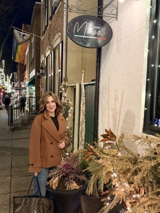 a Woman in a Brown Coat Stands Smiling on a Lit City Sidewalk at Night Beside Winter Greenery Behind Her a Cucina Massi Frederick Md Sign Glows As Festive Lights and Rainbow Flags Decorate the Welcoming Street
