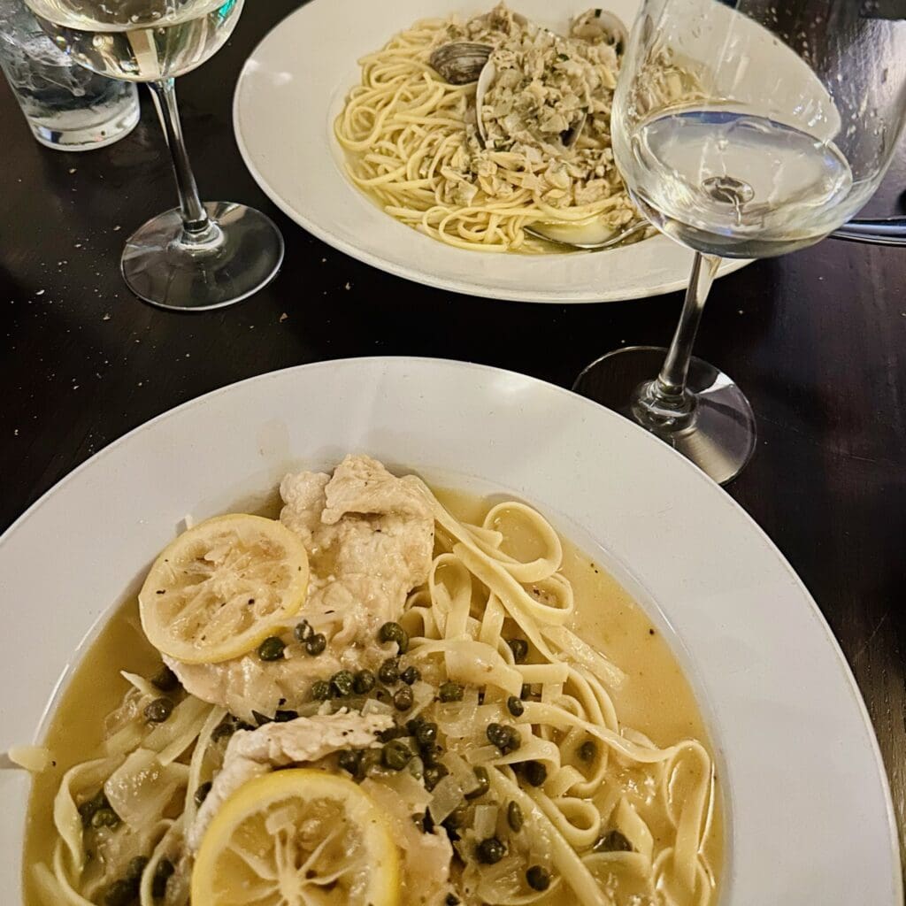 Two Plates of Pasta on a Dark Table at Cucina Massi Frederick Md Front Dish Features Linguine with Lemon Chicken Capers and Sauce Back Dish Has Linguine with Clams Two Glasses of White Wine and a Glass of Water Are Also Visible