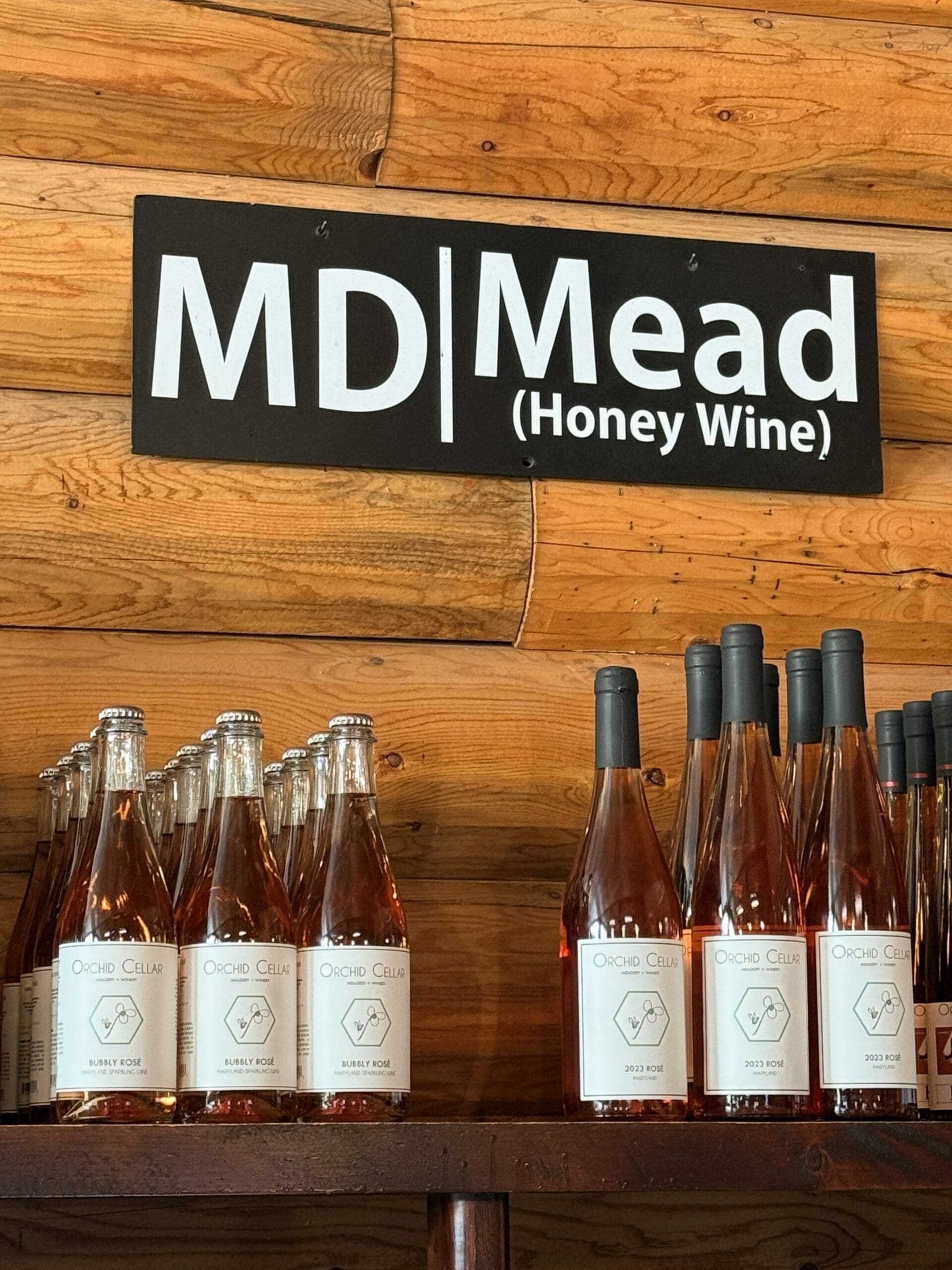 Bottles of Orchid Cellar Mead with White Labels Sit on a Wooden Shelf Below a Black and White Sign Reading md | Mead honey Wine Capturing the Rustic Cozy Charm of Orchid Cellar Middletown Mds Tasting Room