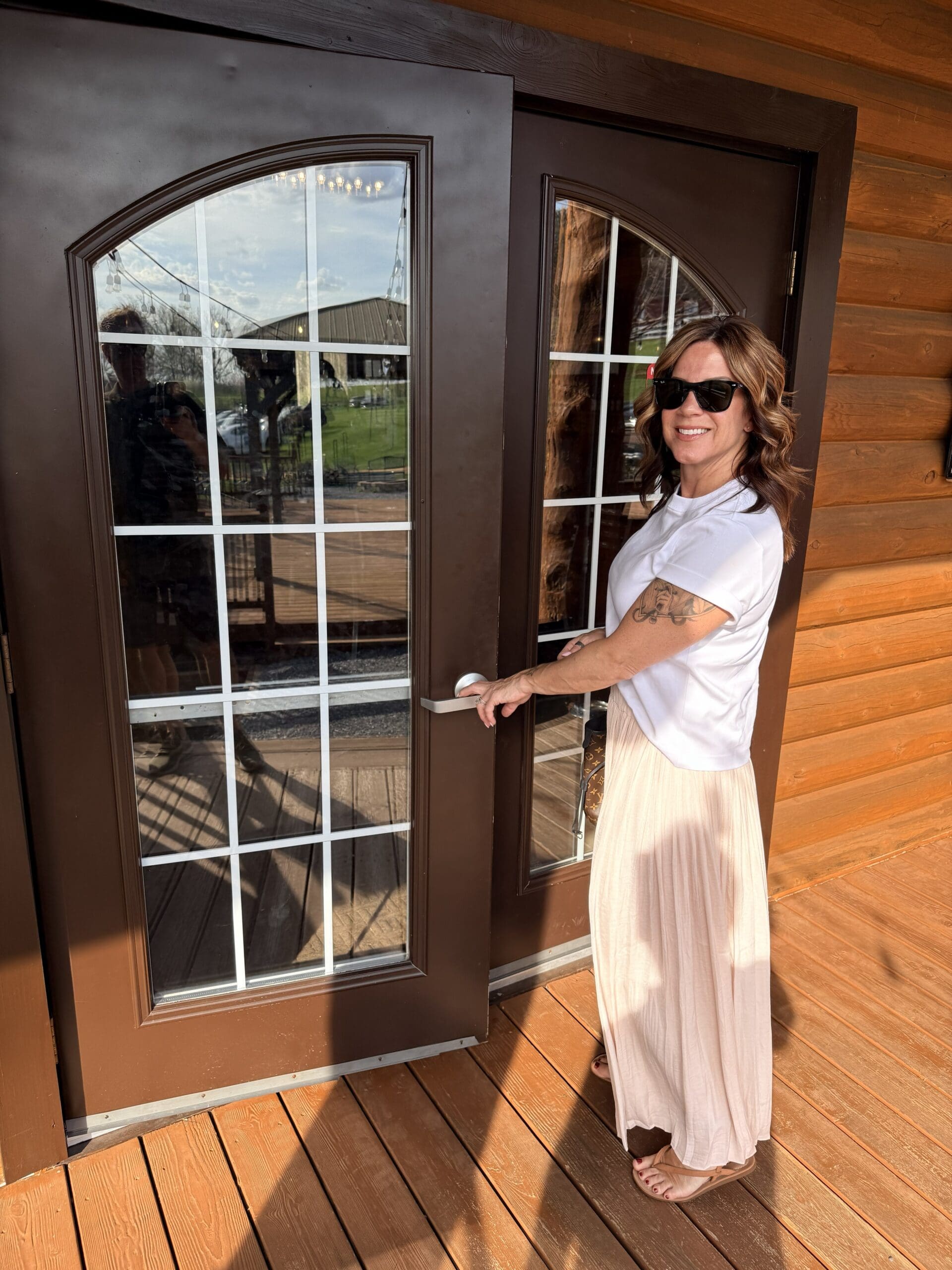 a Woman with Wavy Brown Hair Sunglasses and a White Shirt with a Long Light Pink Skirt Smiles While Opening a Brown Double Glass Door of a Log Cabin at Orchid Cellar Middletown Md Sunlight Creates Reflections on the Door and Wooden Deck