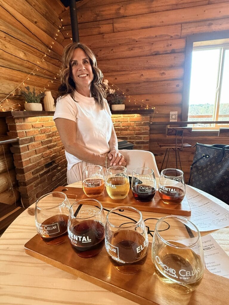 a Woman with Wavy Brown Hair Dressed in White Sits at a Wooden Table in a Cozy Log Cabin at Orchid Cellar Middletown Md Eight Glasses of Mead Each Labeled with a Different Flavor Rest on Trays As Warm String Lights and a Brick Fireplace Glow Behind Her