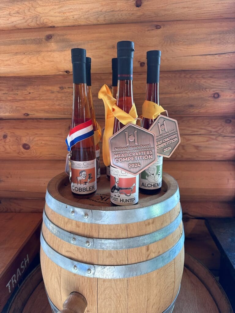 Four Bottles of Orchid Cellar Mead Each Adorned with Medals Rest on a Small Barrel Labeled cobbler frost Hunter and alchemist Theyre Set Against Wooden Walls with a Trash Bin to the Left at Orchid Cellar Middletown Md