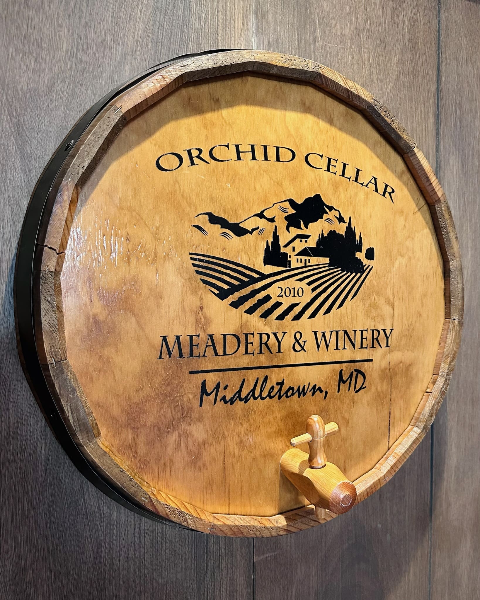 a Round Wooden Barrel Lid with a Spout Mounted on a Wood Wall Displays orchid Cellar Middletown Md 2010 and a Logo of Hills a House Vineyards and Birdsshowcasing the Unique Spirit of Orchid Cellar