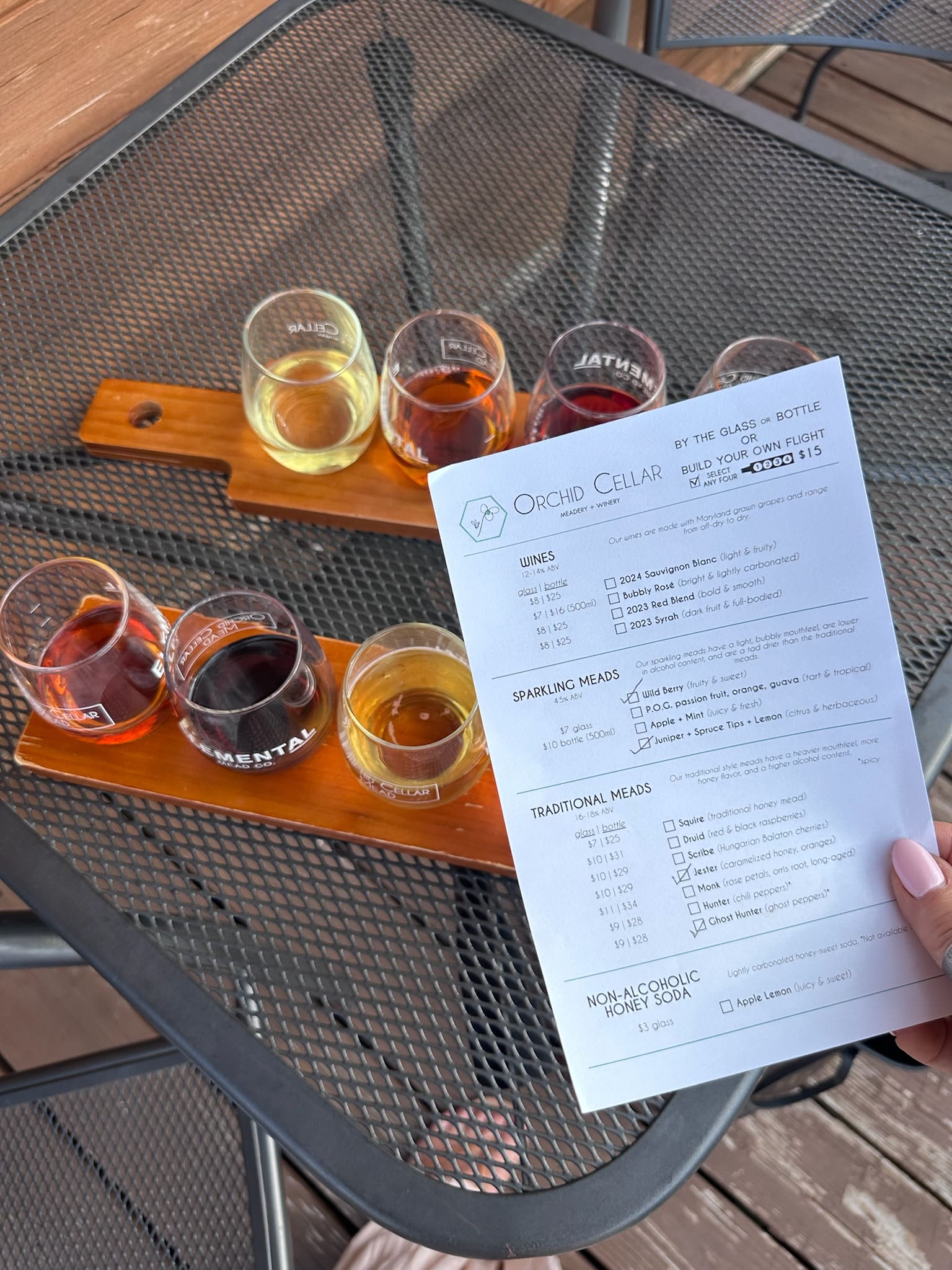 a Hand Holds a Tasting Menu Above Two Wooden Trays Each with Four Small Glasses of Dark Amber and Golden Meads from Orchid Cellar Middletown Md the Trays Rest on a Black Metal Mesh Table Outdoors Sunlight Casting Shadows Across the Surface
