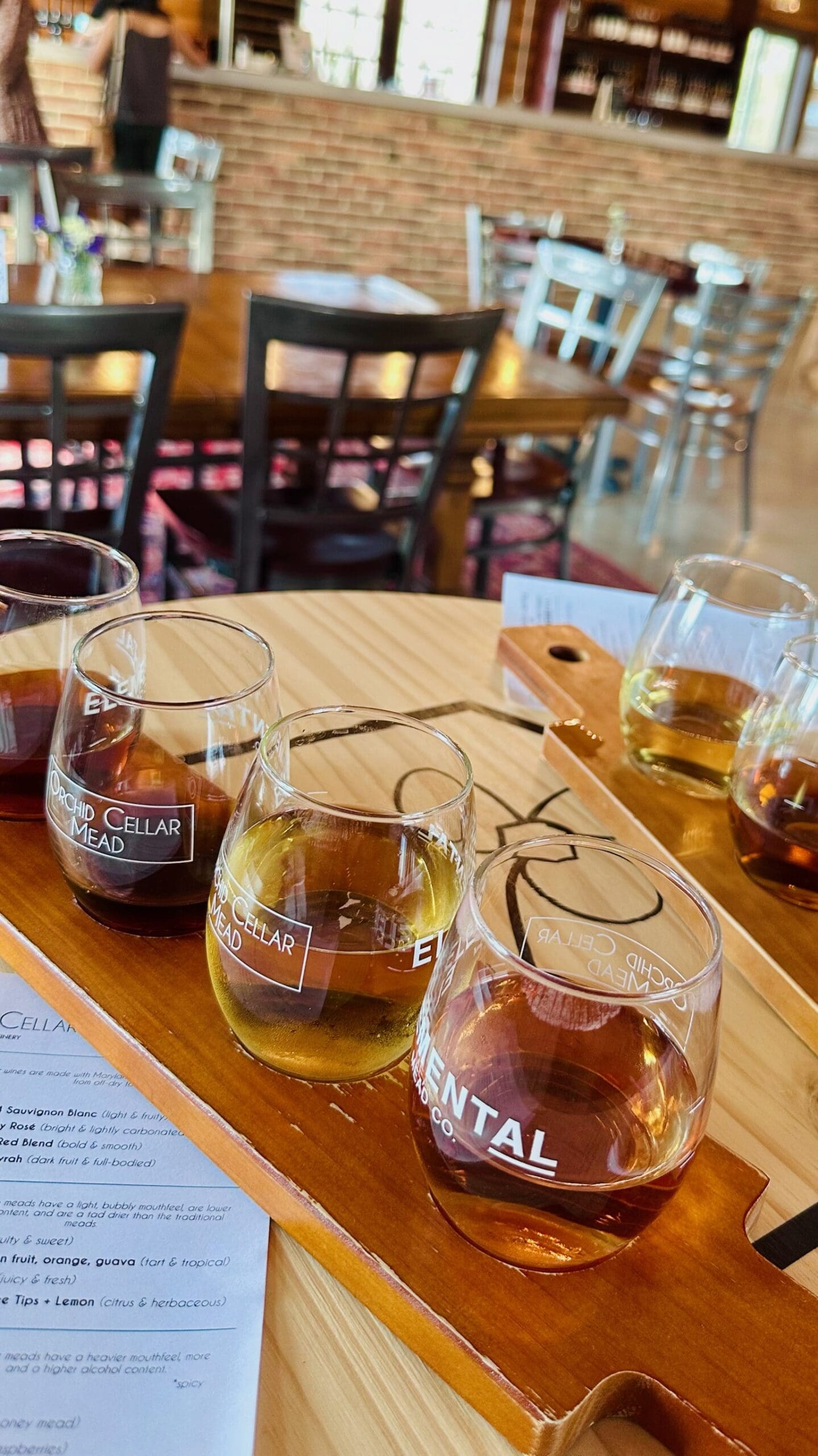 a Wooden Table at Orchid Cellar Middletown Md Holds a Flight of Four Glasses Filled with Different Colored Meads Ranging from Amber to Golden the Glasses Sit on a Labeled Tray with Empty Chairs and a Brick Wall in the Cozy Tasting Room