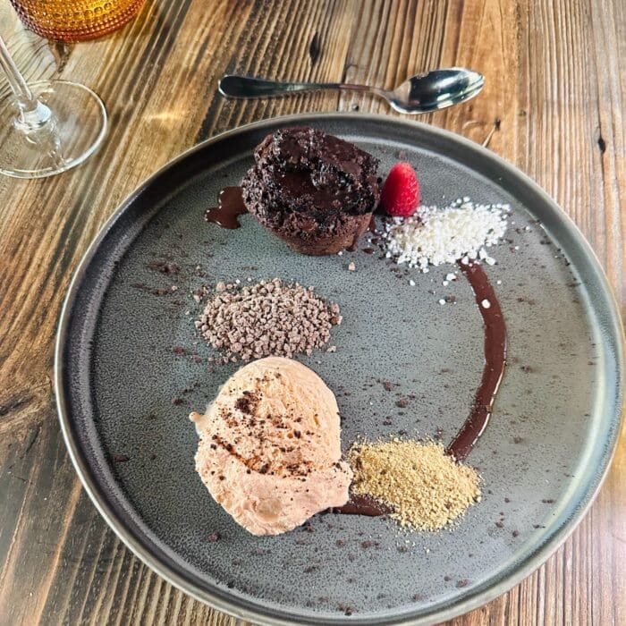 Plate with a Chocolate Dessert Cocoa Powder and a Raspberry on a Rustic Wooden Table with a Spoon Nearby and a Glass at the Edge