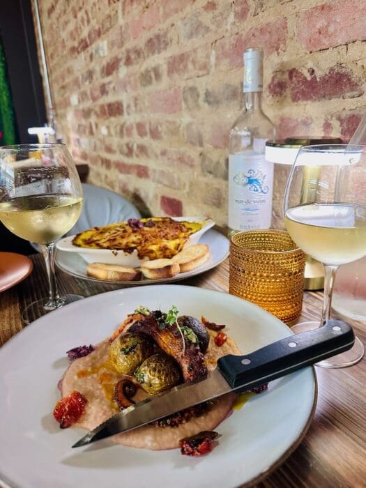 Two Wine Glasses on a Wooden Table with a Bottle of White Wine and Plates of Food Against a Brick Wall Backdrop Suggesting a Relaxed Dinner Setting