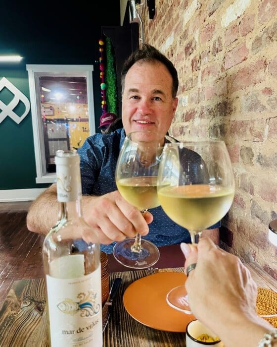 Two People Toasting with White Wine Glasses at a Rustic Brick walled Restaurant a Wine Bottle on the Table Nearby