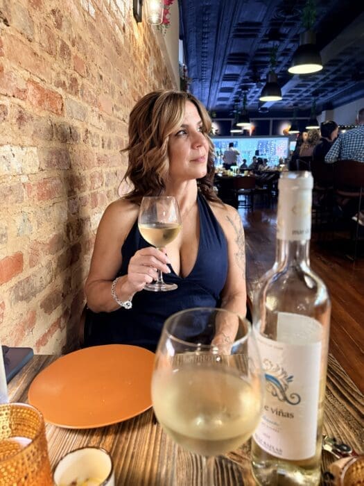 Woman in a Navy Dress Sits at a Wooden Table in a Restaurant Holding a Glass of White Wine with a Brick Wall Behind Her