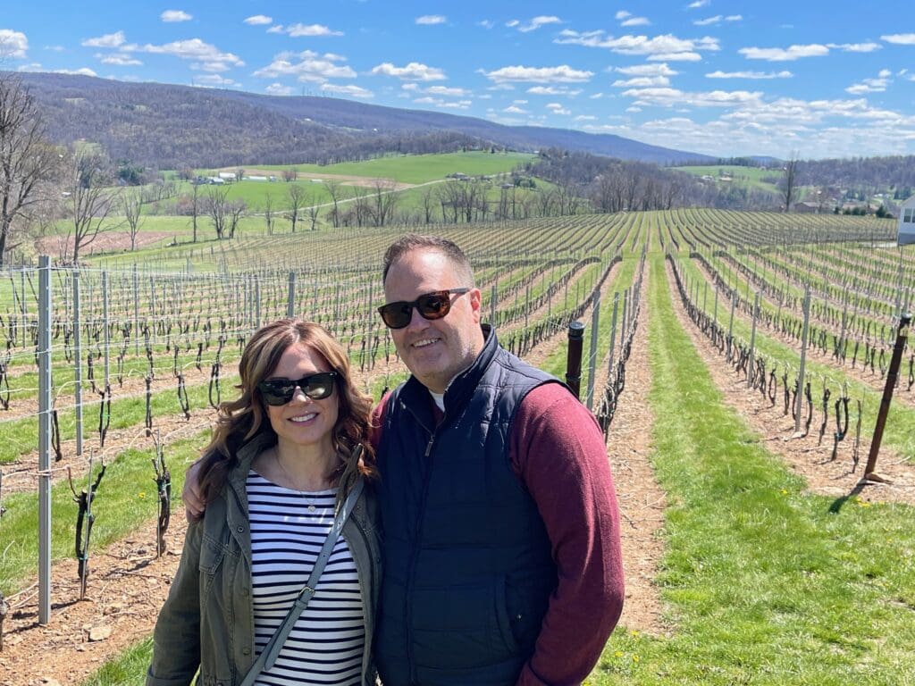 Smiling Couple Posing in a Vineyard with Evenly Spaced Grapevines and Green Hills in the Background Under a Blue Sky