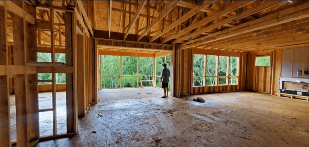 Interior of a House Under Construction with Exposed Wood Framing a Person Looks out over Trees Through Large Windows