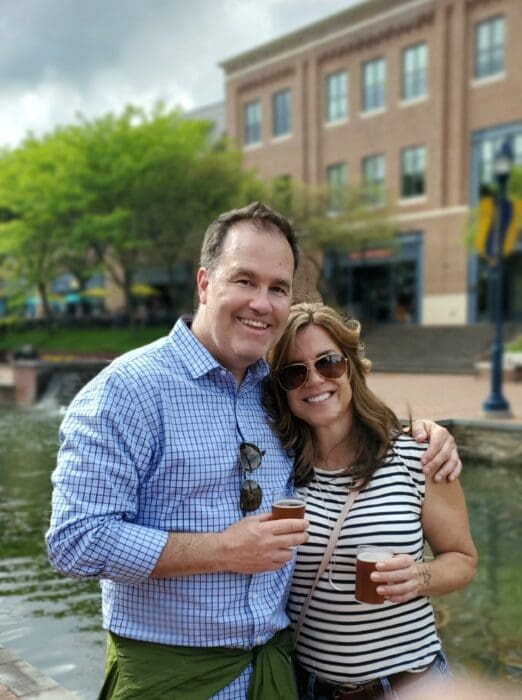 Smiling Couple Posing Outdoors by a Canal with Brick Buildings and Green Trees in the Background Each Holding a Beer Glass