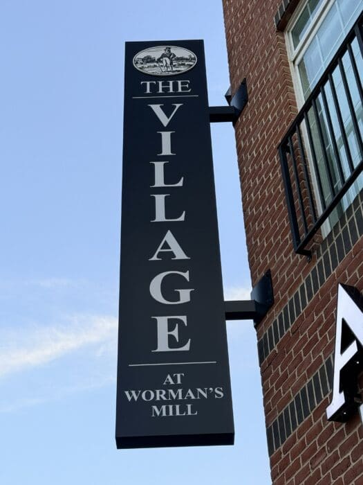 Vertical Black Sign Reading aposthe Village at Wormanaposs Millapos with a Small Oval Logo of a Man in a Landscape Mounted on a Brick Wall in Frederick Md
