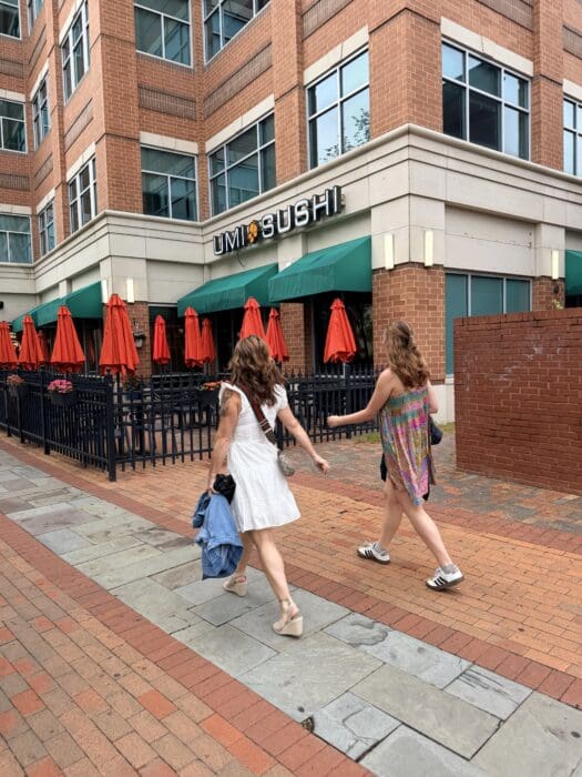 Two Women Walk Past a Brick Umi Sushi Restaurant with Outdoor Seating and Red Umbrellas in Frederick Mda City Known for Its Charming Downtown and Manageable Cost of Living One Wears a White Dress and Holds a Denim Jacket the Other Sports a Colorful Dress and White Sneakers the Sidewalk is Paved with Stone and Brick Reflecting the Inviting Atmosphere That Makes Frederick Md Both Lively and Affordable