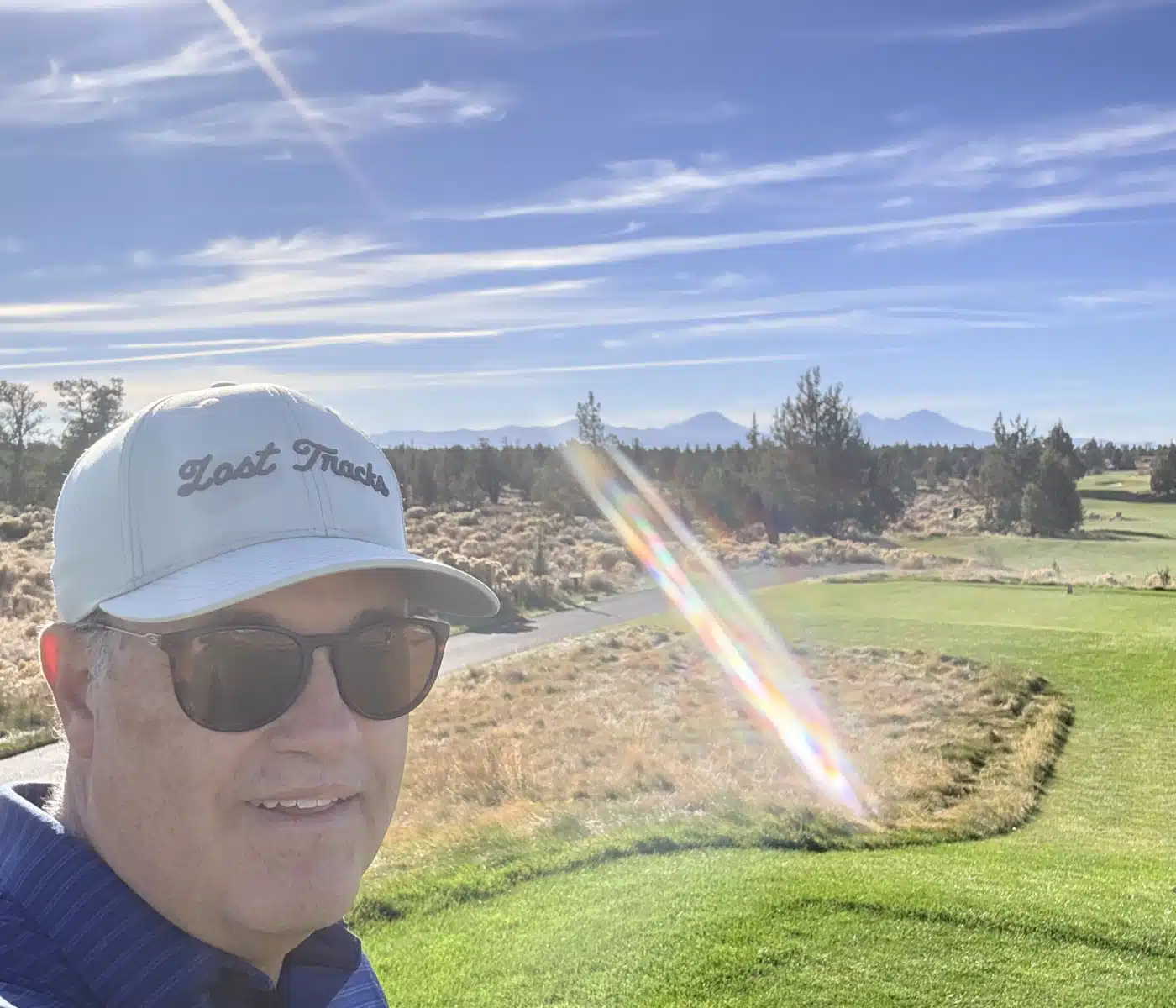 Selfie of a Man Wearing Sunglasses and a Cap That Reads Lost Tracks on a Sunny Day with a Green Golf Course and Distant Mountains in the Background