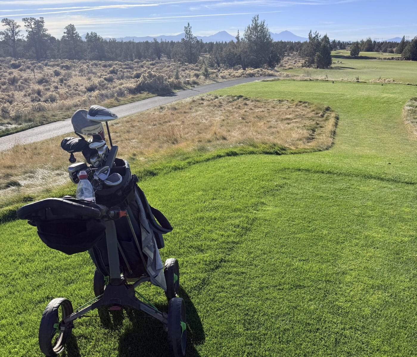 Golf Bag with Clubs on a Cart Beside a Green Fairway Under a Clear Blue Sky Distant Mountains on the Horizon
