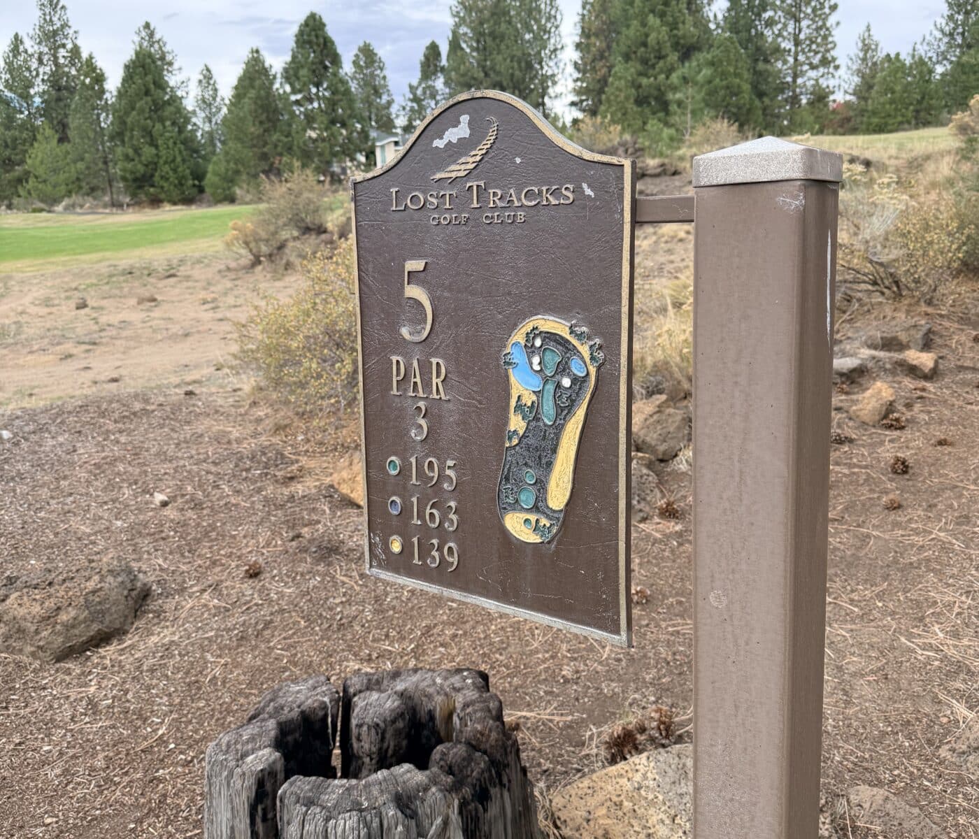 Golf Club Sign Reading Lost Tracks with Hole 5 Par 3 and a Colorful Footprint Graphic Mounted on a Wooden Post Against a Desert Landscape