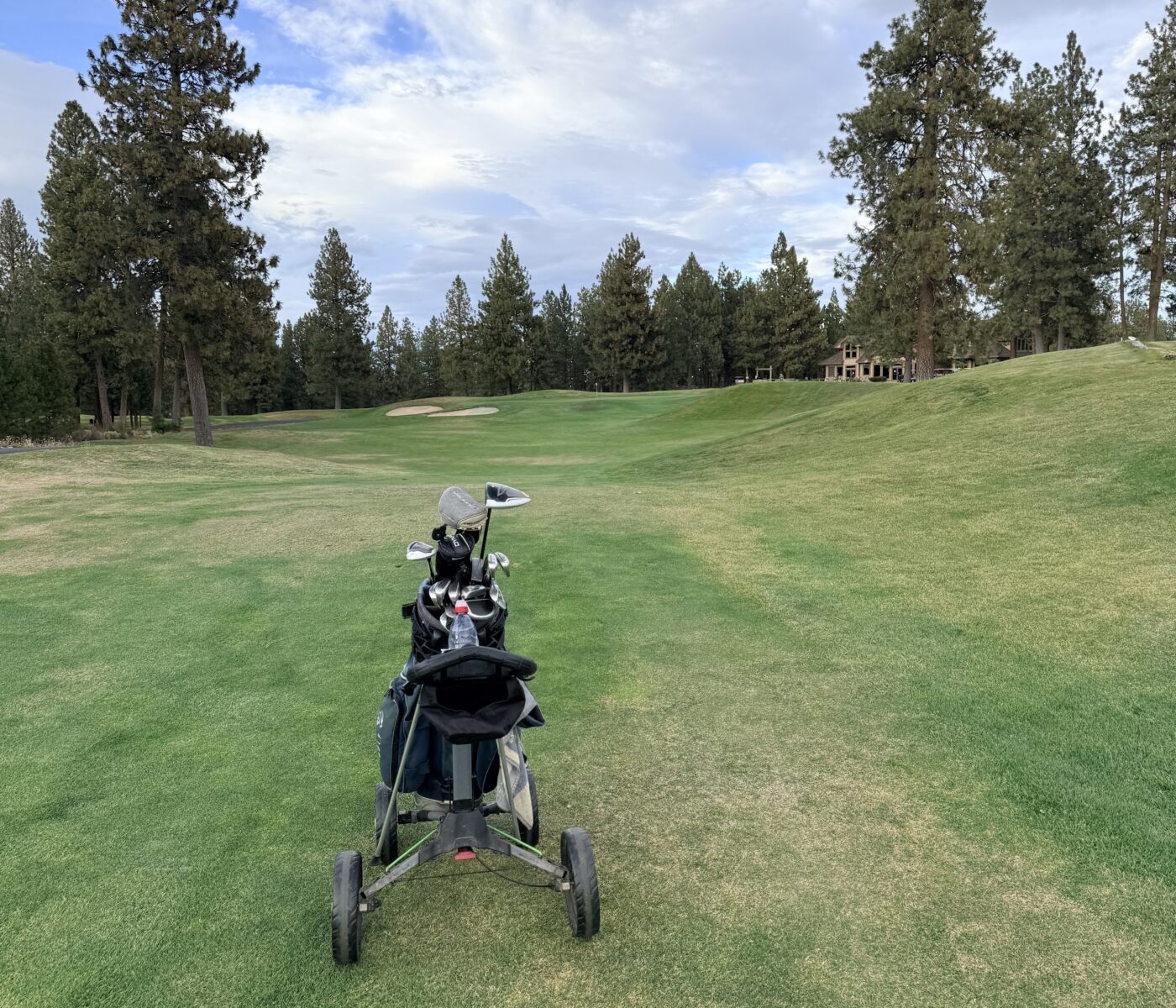 Golf Bag on a Push Cart on a Lush Green Golf Fairway with Tall Trees and a Distant Clubhouse Under a Partly Cloudy Sky