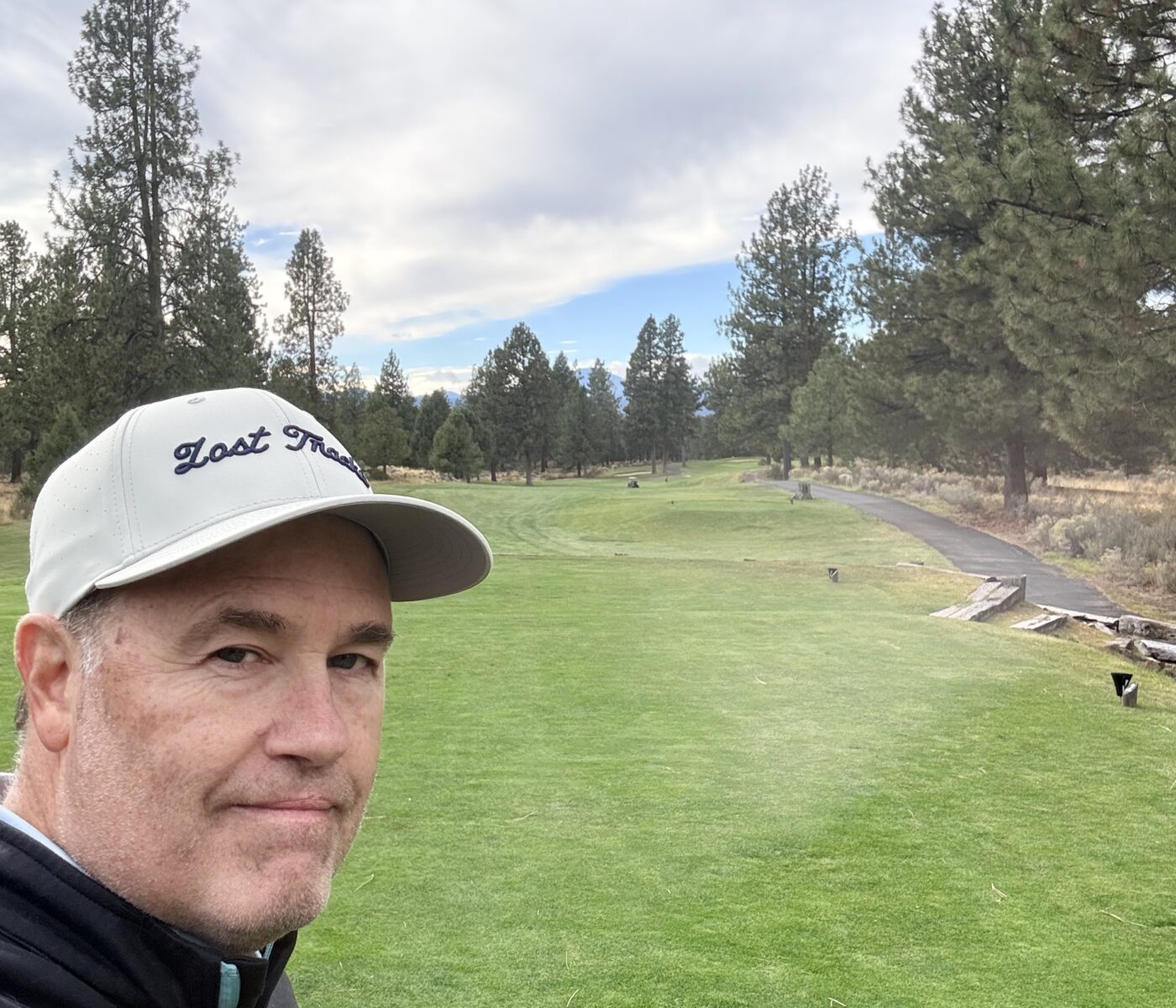 Selfie of a Man Wearing a White Cap on a Green Golf Course with Tall Trees and Cloudy Sky in the Background