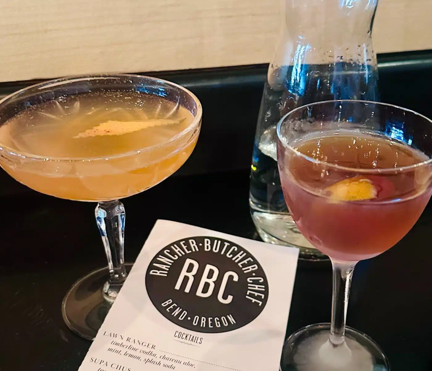 Two Cocktails in Stem Glasses with a Carafe on a Dark Bar and a Rbc Bend Oregon Menu in the Foreground