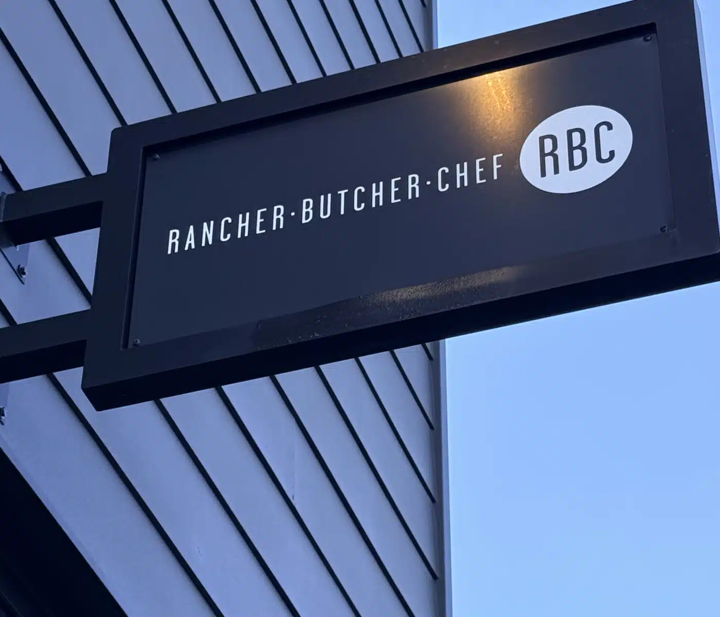 Black Rectangular Hanging Sign on a Building Reading Rancher  Chef with an Rbc Oval Logo Set Against a Blue Sky