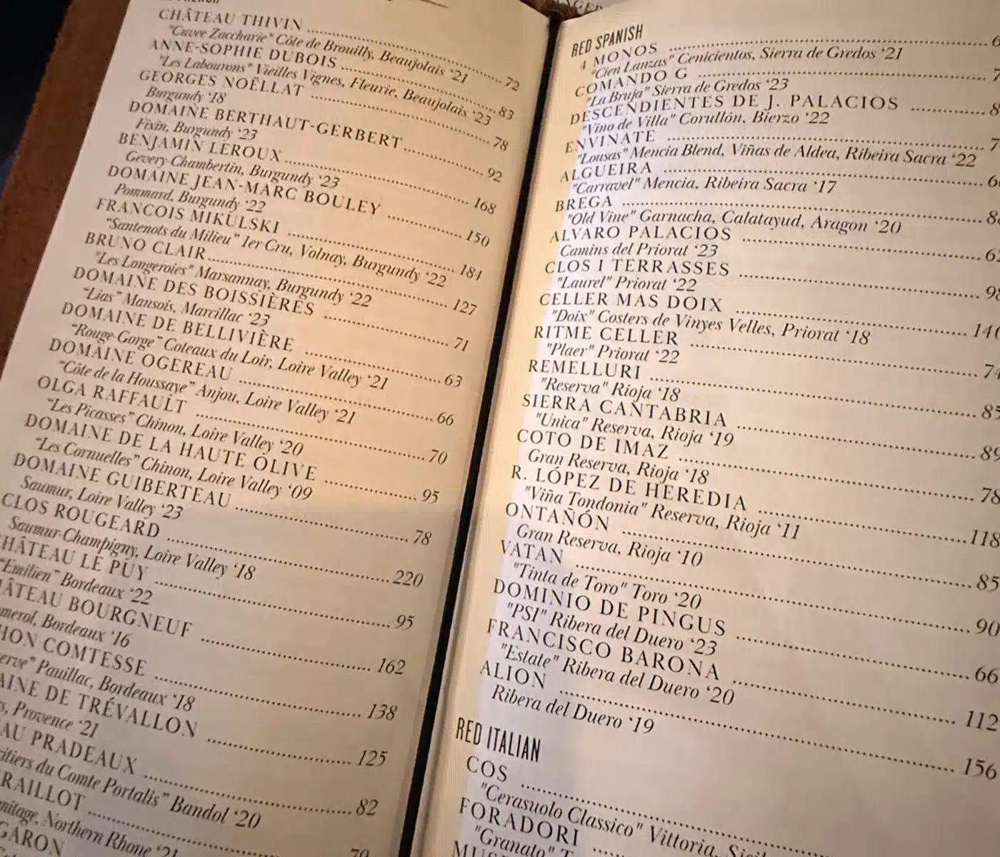 Open Wine Menu Laid Flat on a Table Showing Old World Red on the Left Page and Red Spanish on the Right with Small Print Wine Entries and Dotted Separators a Round Rbc Bend Oregon Badge is Visible in the Background