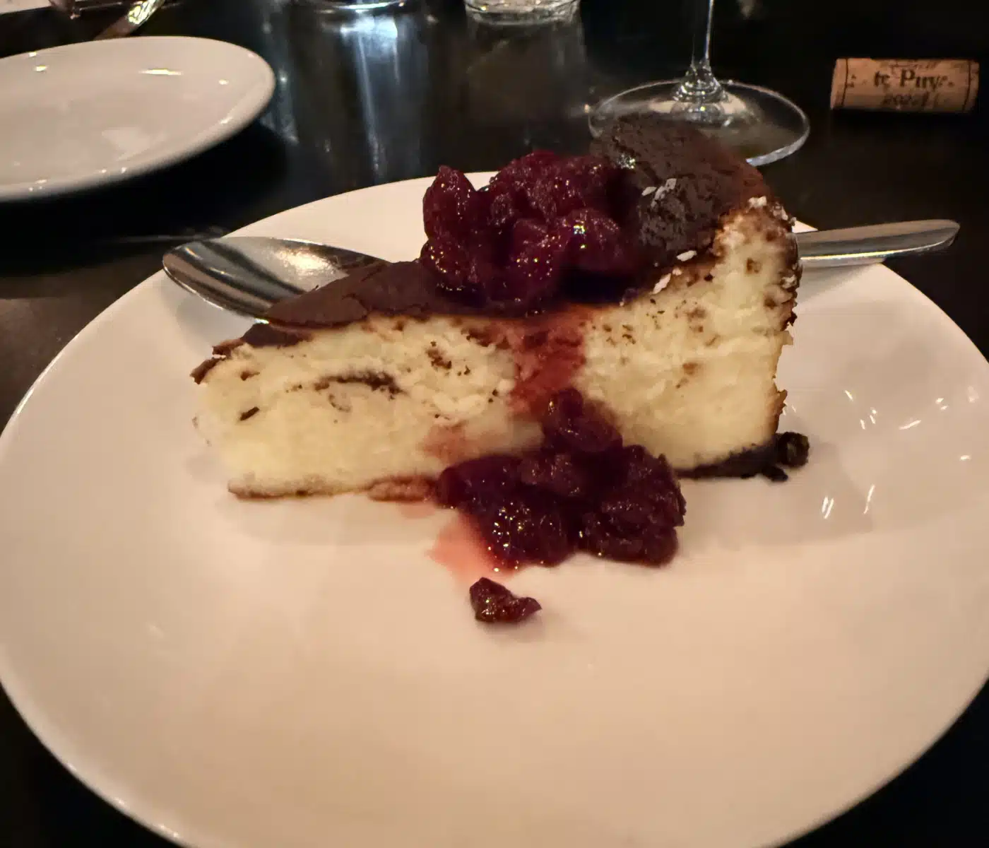 a Slice of Vanilla Cake with Cherry Topping on a White Plate Knife and Spoon Nearby with a Wine Glass in Background