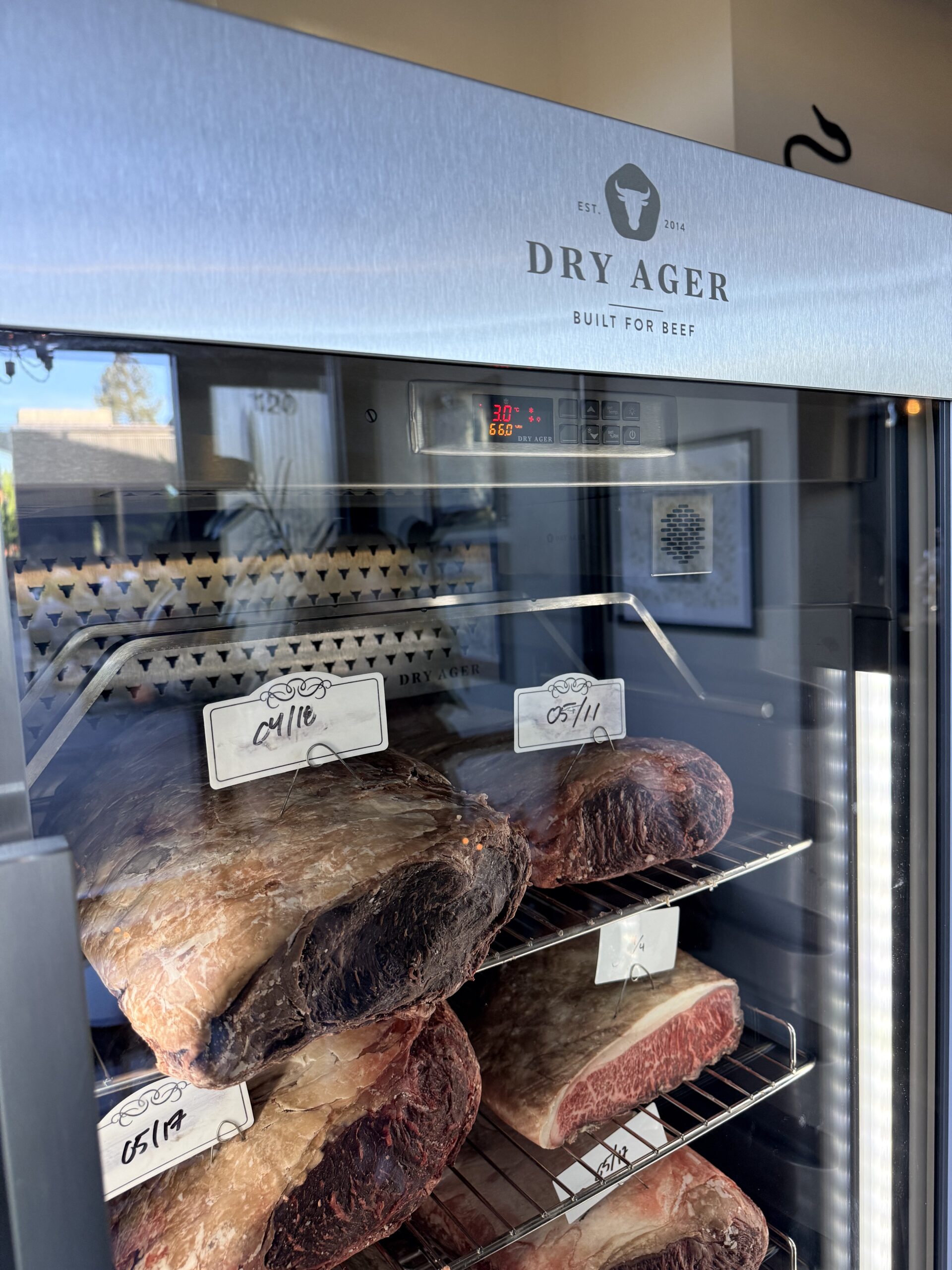 Display Case with Large Dry Aged Beef Cuts on Metal Racks Inside a Glass Front Refrigerator Logo Reads Dry Ager with Built for Beef Tagline