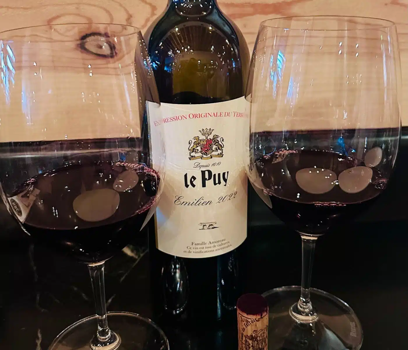 Bottle of Red Wine Centered Between Two Filled Wine Glasses on a Dark Surface Cork to the Side Label Partially Visible Reading Le Pu Emilien