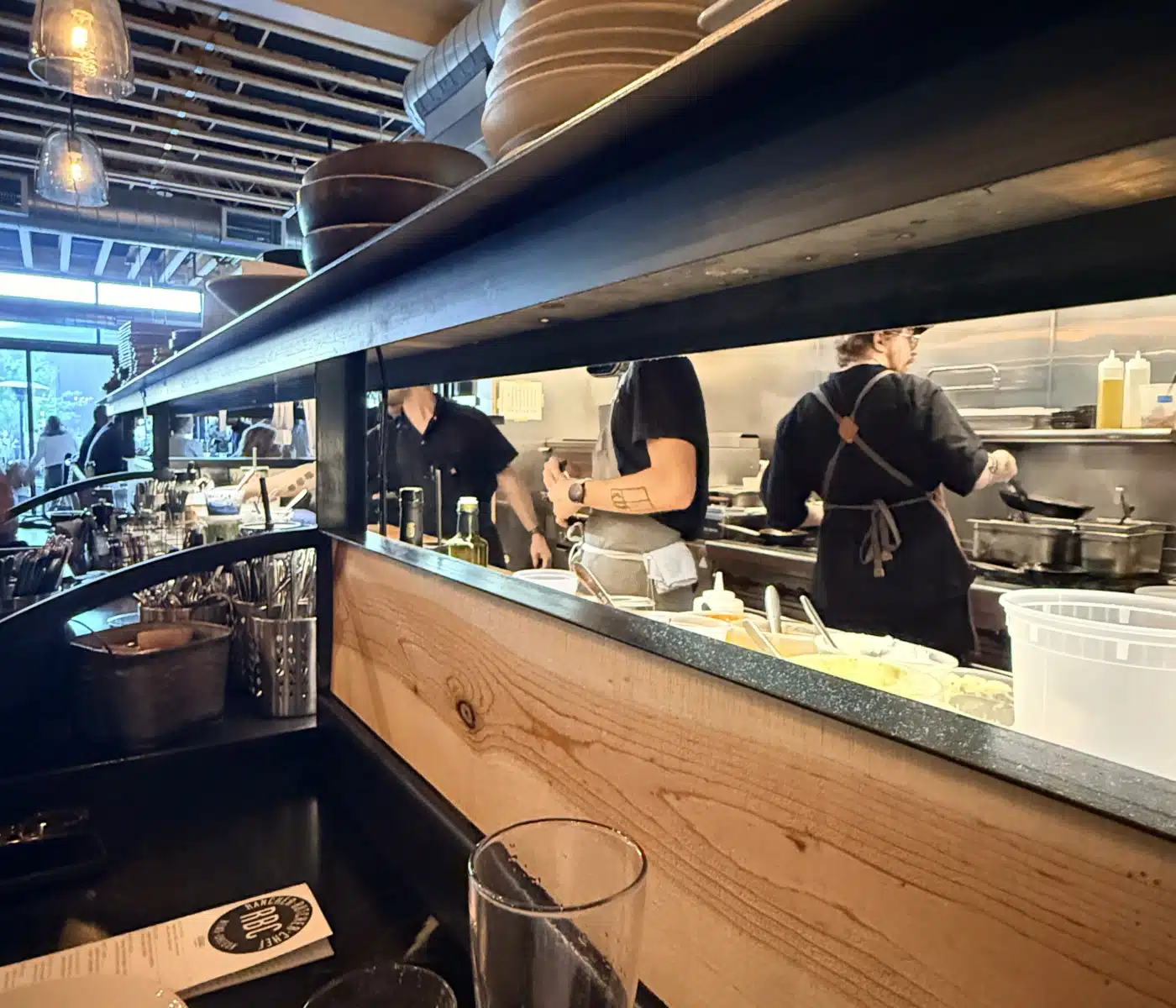 Open Kitchen Restaurant Scene with Staff Cooking Behind a Counter Stacked Plates on a Shelf and Hanging Glass Lamps Above the Prep Area