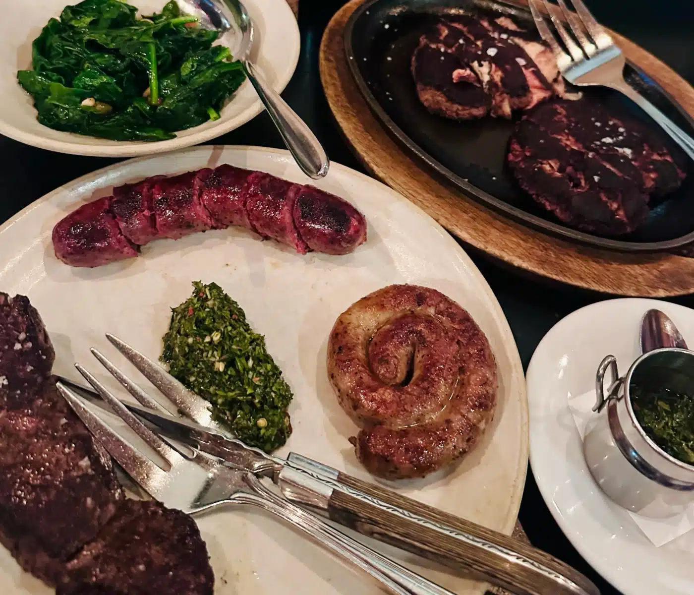 Plate with Several Cooked Beef Steaks and a Side of Sautéed Greens Plus a Skillet of Meat and Cutlery Nearby