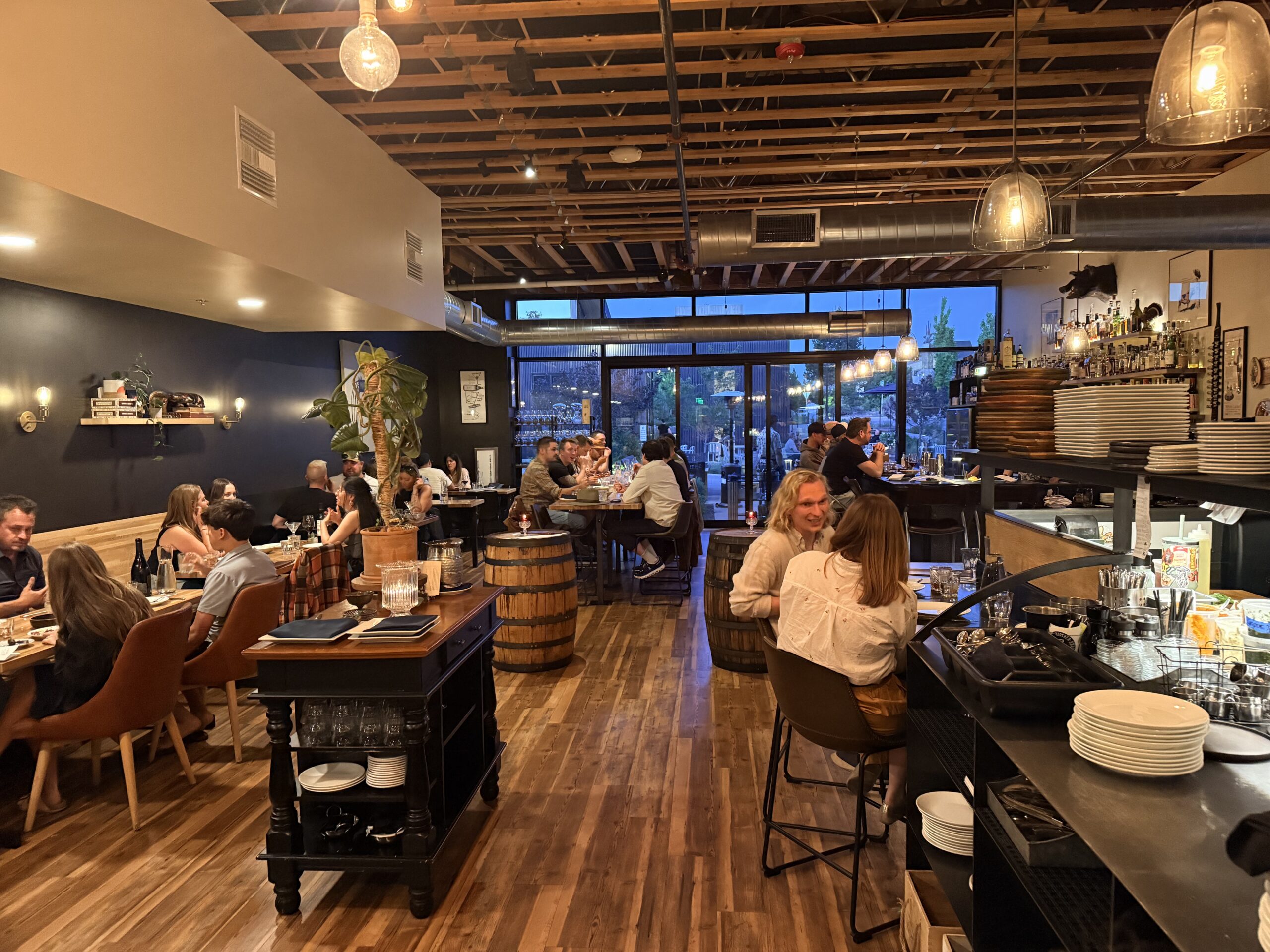 Bustling Modern Restaurant Interior with Diners at Tables Warm Pendant Lighting and an Open Bar Area to the Right dining Room with Wooden Floors Exposed Ceiling Beams and Groups of People Enjoying Meals at Long Tables restaurant Scene at Dusk with Busy Seating Bar Shelves and Industrial style Lighting customers Eating at Long Wooden Tables in a Stylish Open plan Dining Space with a Visible Kitchenbar Area