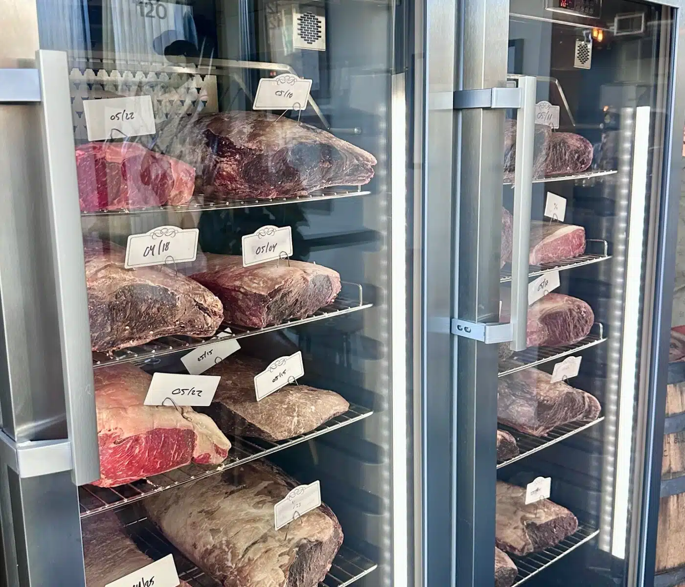 Glass Door Meat Aging Display with Large Beef Cuts and Handwritten Price Tags in a Dry Ager Cabinet
