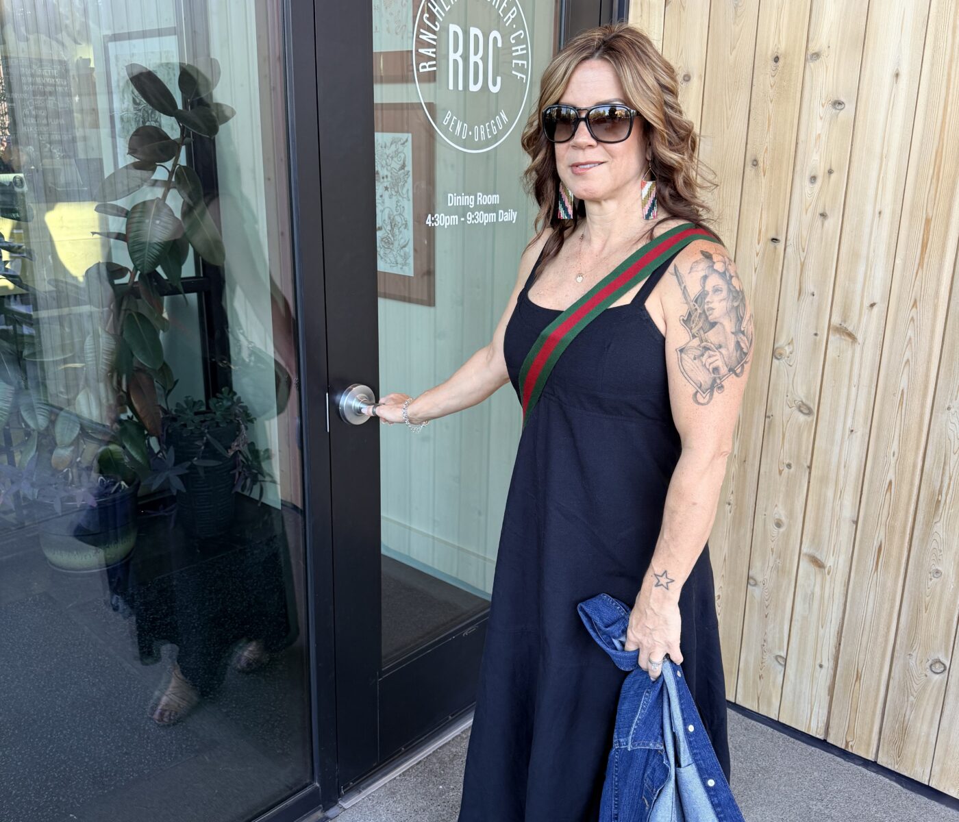Woman in a Black Sundress with a Green Red Strap Across Her Chest Stands by a Glass Door Holding a Denim Jacket and Wearing Sunglasses Tattoo Sleeve Visible on Her Left Arm