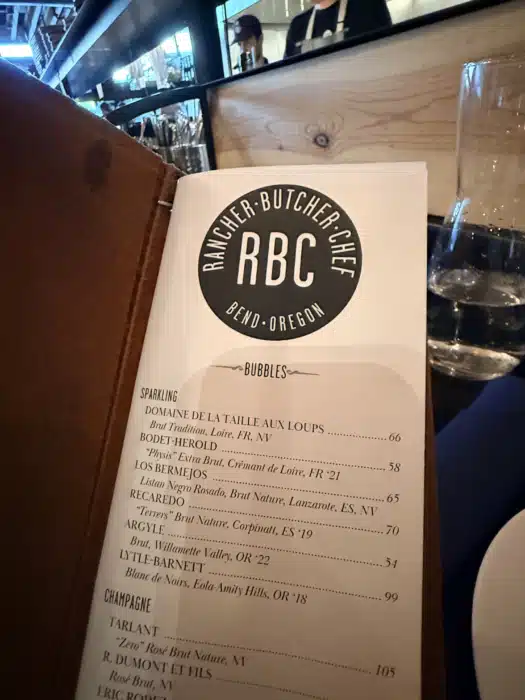 Menu Page with a Large Circular Rbc Logo rbc Bend Oregon Visible Resting on a Wooden Surface with a Leather Edge Nearby the Page Includes a Section Titled bubbles and Lists Sparkling Wines