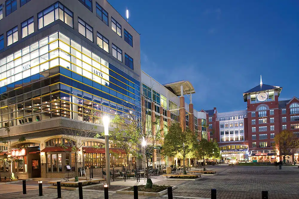 Evening Shopping Plaza with Glass Storefronts Outdoor Seating and a Red Brick Clock Tower Building in the Distance