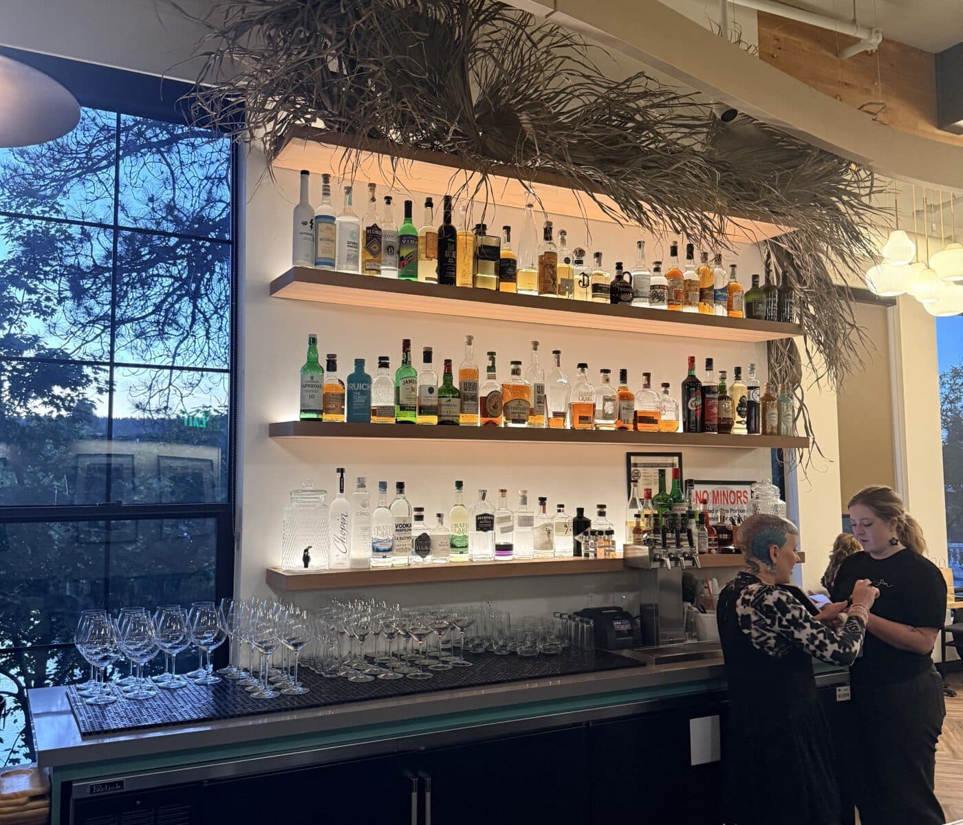 Two Bartenders Work Behind the Sen Thai Noodles Bar Which Features Three Backlit Shelves Lined with Diverse Liquor Bottles and a Large Arrangement of Dried Grasses Overhead to the Left a Large Window Overlooks a Dark Outdoor Scene While Rows of Empty Wine Glasses Sit on the Counter