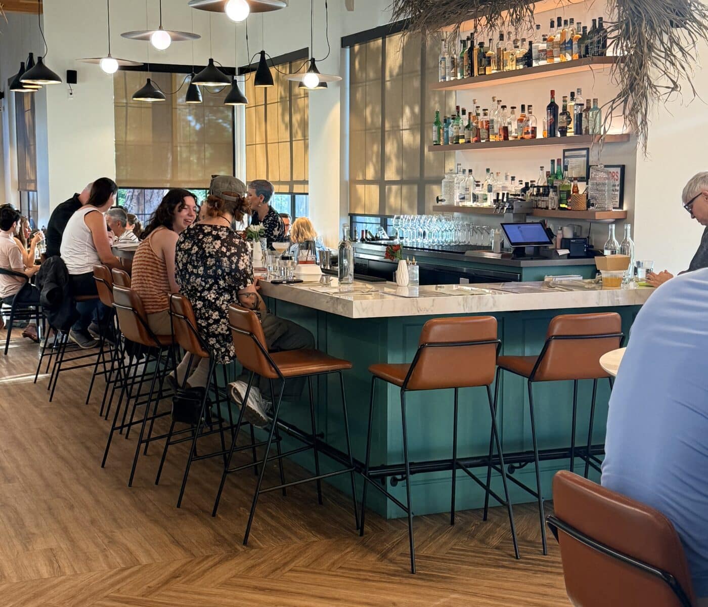 Patrons Sit at a Teal Bar Counter with a White Marble Top at Sen Thai Noodles the Bright Modern Restaurant Features Rows of Glass Bottles on Shelves Leather Stools and Distinctive Curved Lighting Fixtures Suspended from the Ceiling